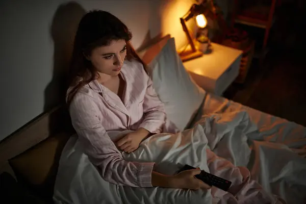 Una donna si siede sul suo letto, avvolta in coperte, fissando uno schermo mentre si sente isolata. — Foto stock