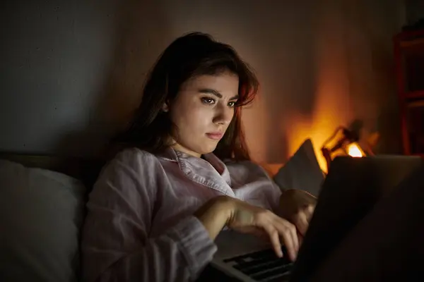In a dimly lit room, a woman sits on her bed, deeply absorbed in her laptop, wrestling with sadness. — Stock Photo