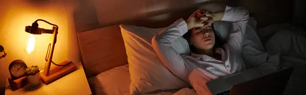 Una donna si siede a letto, mano sulla fronte, persa nel pensiero sotto una luce soffusa. — Foto stock