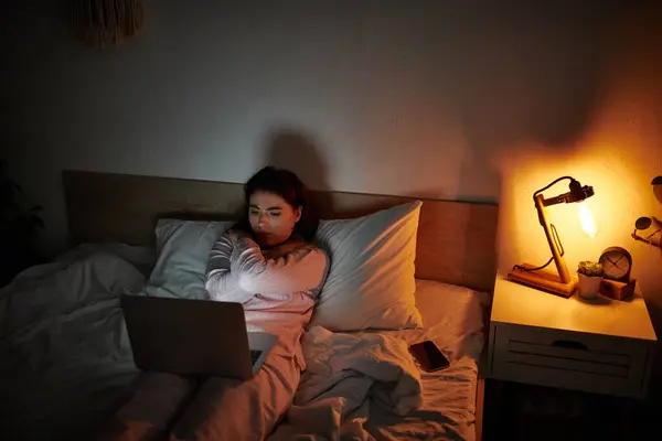 A woman sits alone in bed, illuminated by a soft lamp, lost in deep thoughts at night. — Stock Photo