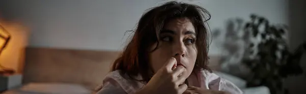 A troubled woman sits on her bed, grappling with her emotions in a dimly lit room. — Stock Photo