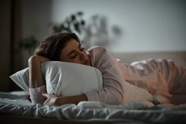 Una mujer yace de costado en la cama, agarrando una almohada mientras experimenta emociones dolorosas. - foto de stock