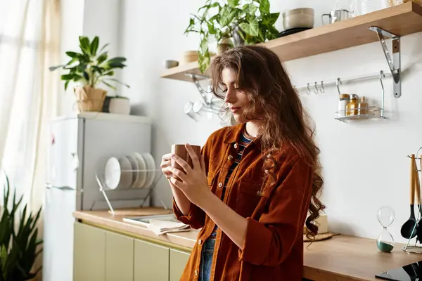 Une femme se tient pensivement dans sa cuisine, tenant une tasse chaude près, perdue dans ses émotions. — Stock Photo