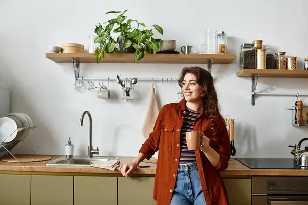 In una cucina calda, una donna si appoggia al bancone, tenendo in mano una tazza, persa nel pensiero. — Foto stock