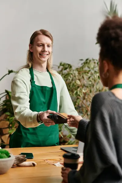 Pretty woman paying with her phone while man working in green setting. — Stock Photo
