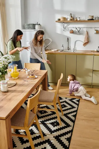 Two women joyfully interact as their young daughter plays on the floor in a stylish kitchen. — Stock Photo
