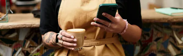 A beautiful young woman holds a steaming cup of coffee while focused on her smartphone. — Stock Photo
