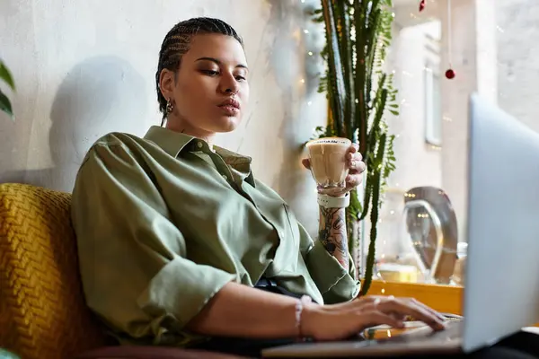 In a vibrant cafe, a stylish young woman savors her coffee while focused on her laptop screen. — Stock Photo