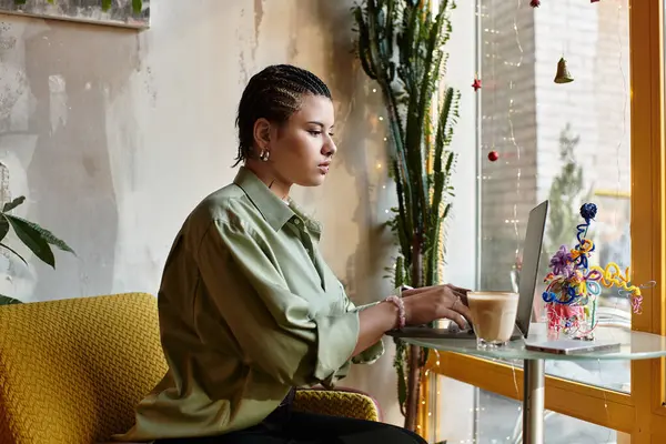 Dans un café confortable, une jeune femme avec une coiffure unique sirote du café et travaille sur son ordinateur portable. — Photo de stock