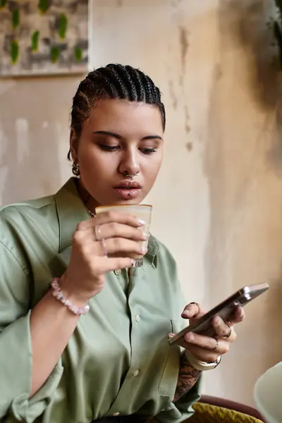 A young woman with unique style sips her drink while focused on her phone at a cozy cafe. — Stock Photo