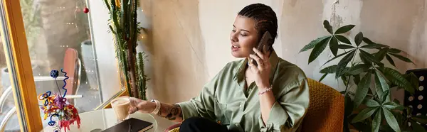 A young woman sips coffee in a bright, green cafe, chatting animatedly on her phone. — Stock Photo