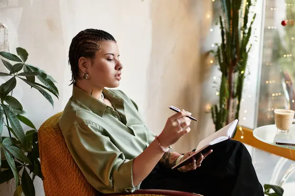 In a vibrant cafe, a young woman pens her thoughts with focus and style, surrounded by greenery. — Stock Photo