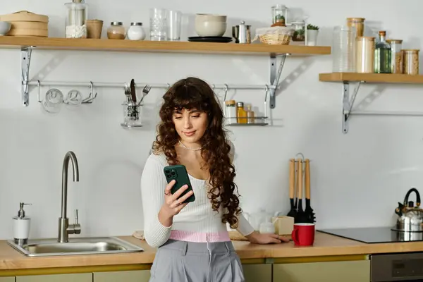 In a bright kitchen, a young woman smiles while happily scrolling her smartphone. — Stock Photo