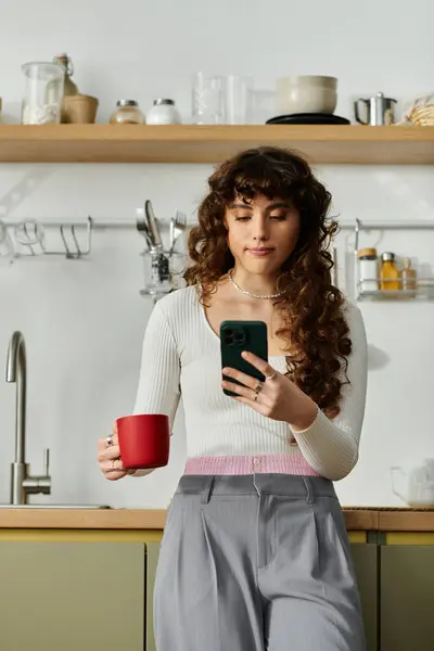 A young woman stands in a sleek kitchen, sipping coffee and browsing her phone, lost in thought. — Stock Photo