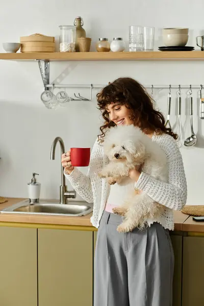 Una joven encantada sostiene una taza roja y un perro esponjoso en una cocina caliente llena de luz. - foto de stock