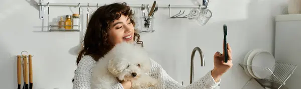 Mujer joven sonriente sostiene a su perrito esponjoso, la captura de selfies alegres en una cocina iluminada por el sol. — Stock Photo