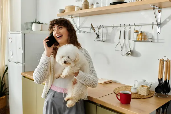 Una joven sonriendo mientras sostiene a un perro esponjoso mientras habla en su teléfono inteligente en una cocina luminosa. - foto de stock