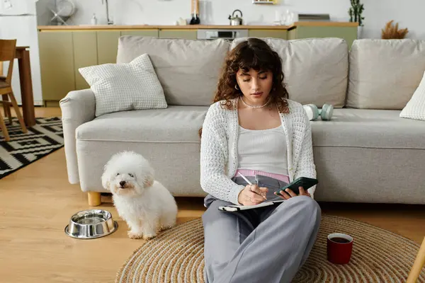 Uma jovem mulher se senta no chão, escrevendo em seu caderno com seu cachorro fofo ao seu lado. — Fotografia de Stock