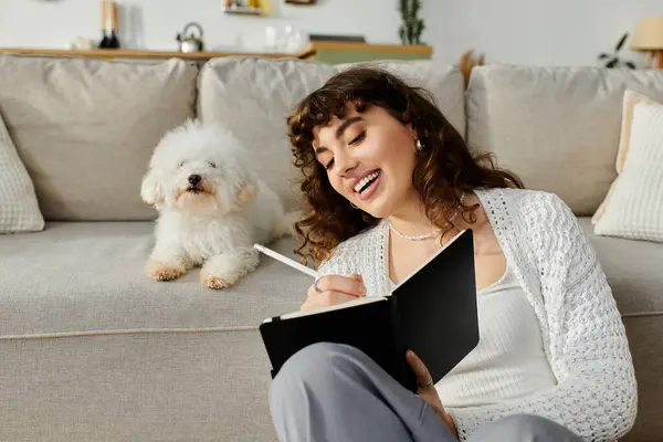 A young woman is smiling while writing in her notebook. Her fluffy dog sits nearby on the couch. — Stock Photo