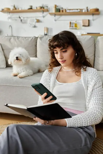A young woman enjoys reading a book while checking her phone in a bright living room with a dog. — Stock Photo