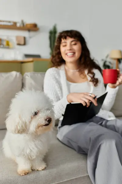 Relaxing on a sofa, a young woman smiles and reads, accompanied by her adorable fluffy companion. — Stock Photo