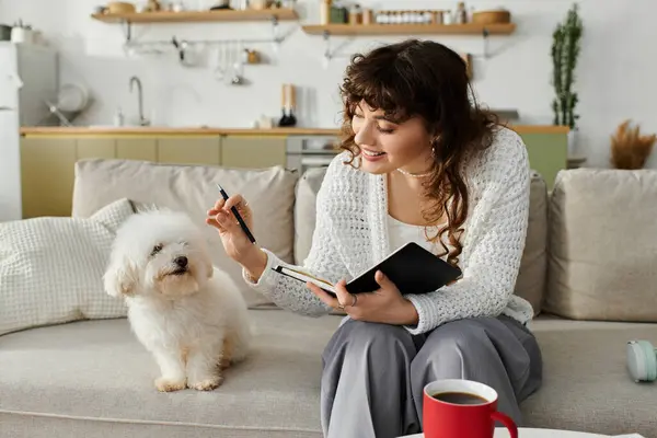 A young woman sits comfortably on a couch, pen in hand, jotting down thoughts with her dog nearby. — Stock Photo
