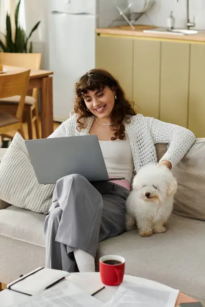 A young woman relaxes on the couch, engaging with her laptop and accompanied by her fluffy dog. — Stock Photo