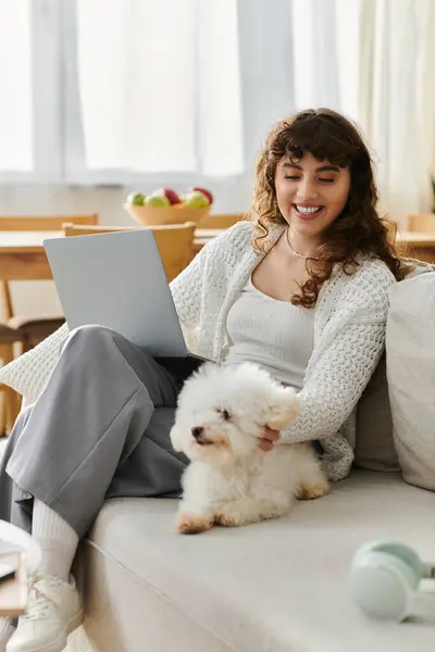 A young woman is happily engaged with her laptop, sitting on a sofa with her dog beside her. — Stock Photo