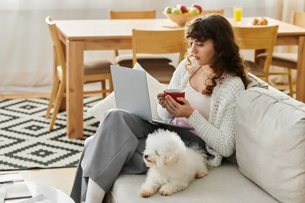 Relaxing in a bright living room, a young woman sips a warm drink and works on her laptop. — Stock Photo