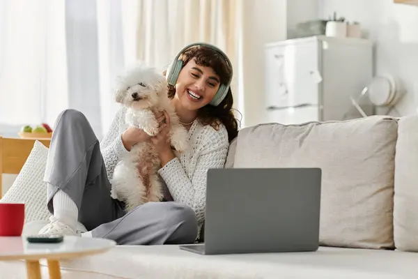 A cheerful young woman sits comfortably on a sofa with her fluffy dog, engaging in a virtual call. — Stock Photo