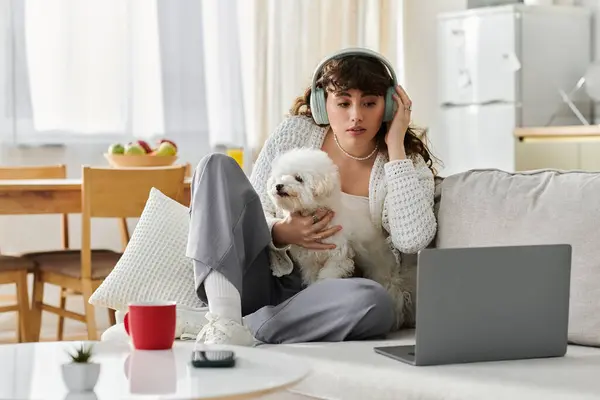 A young woman sits comfortably on a couch, wearing headphones, with her small dog on her lap. — Stock Photo