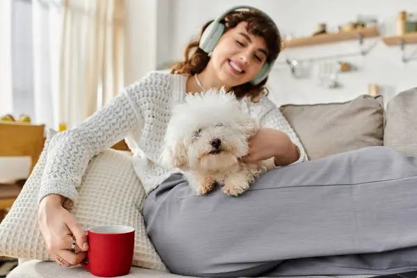 A young woman enjoys her time relaxing on a couch, cuddling a small dog while sipping coffee. — Stock Photo