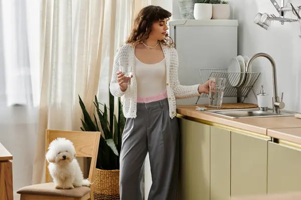 In a bright kitchen, a young woman sips water while her dog curiously watches her. — Stock Photo
