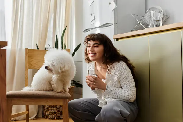 A young woman kneels on the floor, smiling with her drink and fluffy dog. — Stock Photo