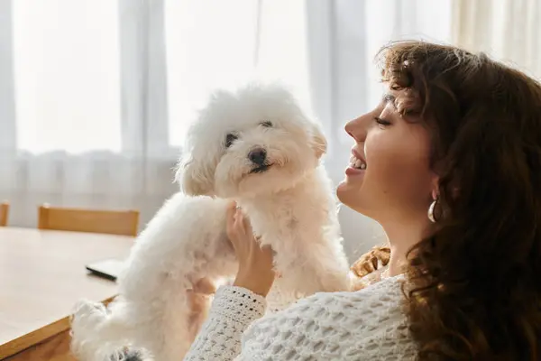 A young woman smiles brightly while holding her fluffy white dog in a cozy, sunlit room. — Stock Photo
