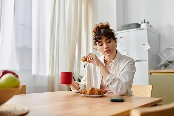 A young woman enjoys a croissant and sips from a red mug at a wooden table. — Stock Photo
