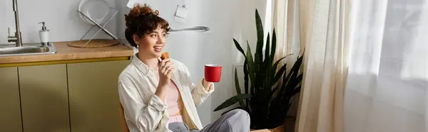 Young woman sits comfortably, sipping coffee and enjoying a snack in a bright indoor space. — Stock Photo