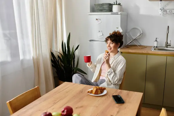 A young woman relaxes in a sunny kitchen with a red cup and pastries. — Stock Photo