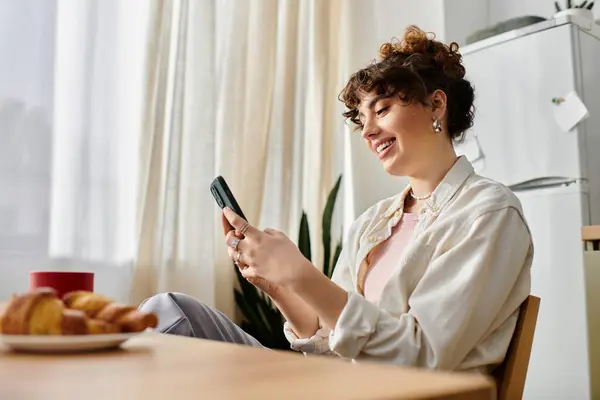 A young woman smiles while texting at a wooden table, surrounded by pastries and warm light. — Stock Photo