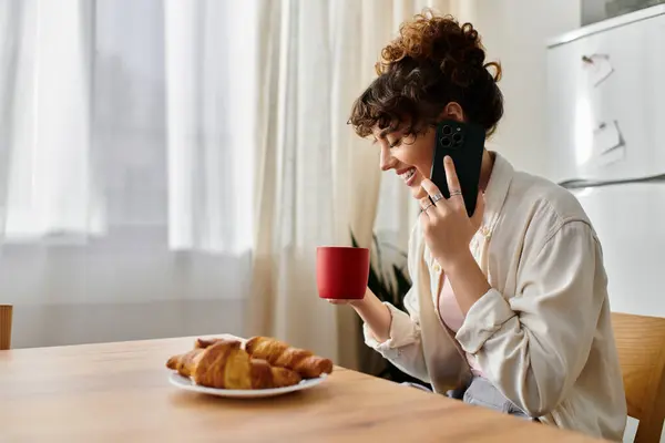 A young woman smiles while sipping coffee and holding a smartphone in a cozy setting. — Stock Photo