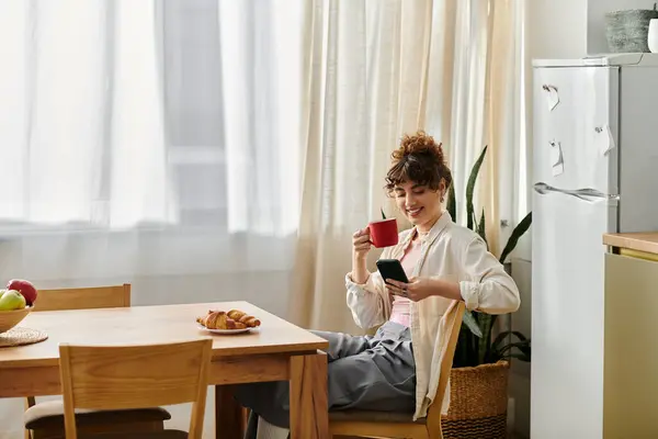 A young woman smiles while sipping coffee and browsing her phone at a sunlit kitchen table. — Stock Photo