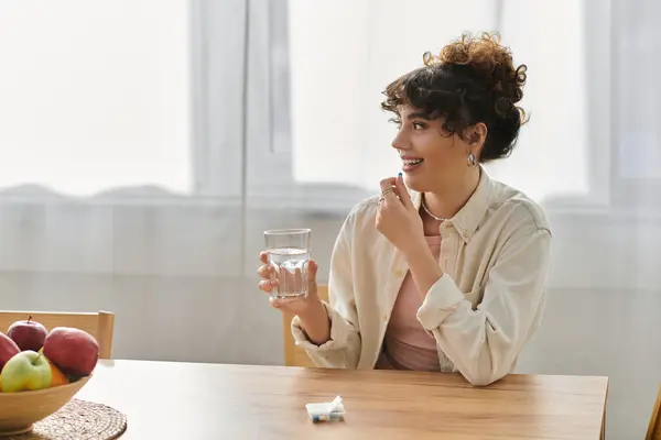 Woman smiles with a glass of water at a wooden table filled with fruit in a cozy kitchen. — Stock Photo