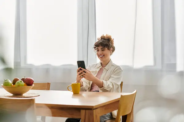 A young woman smiles at her smartphone while sipping coffee at a sunlit kitchen table. — Stock Photo