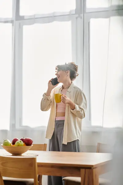 Una joven mujer está parada en una cocina iluminada por el sol, sosteniendo una taza mientras charla felizmente en su teléfono. - foto de stock