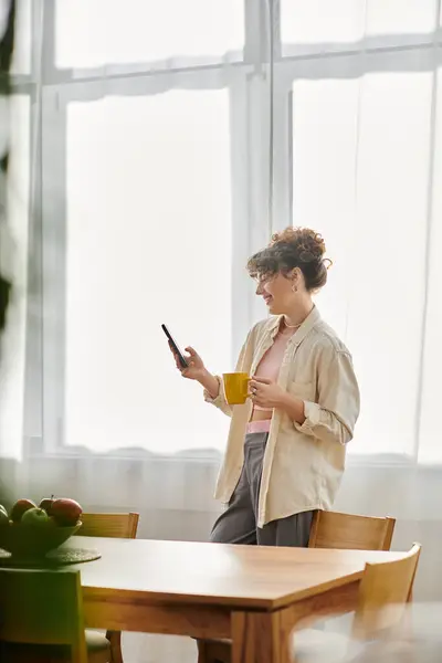 A young woman stands by a table, holding a cup and engaged with her smartphone in soft sunlight. — Stock Photo