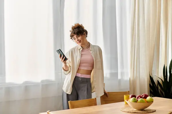 In a bright and airy room, a young woman stands by a wooden table, engaged with her phone. — Stock Photo