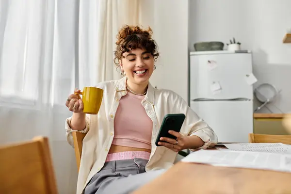 Mujer sonriente sorbe café de una taza amarilla mientras se desplaza en su teléfono en una acogedora mesa de la cocina. - foto de stock