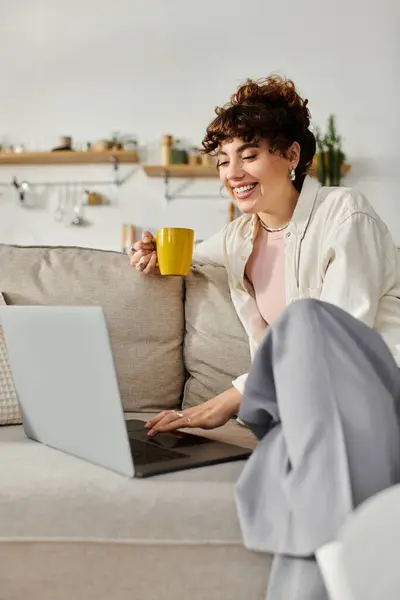 A young woman sits on a sofa, sipping coffee and joyfully focused on her laptop. — Stock Photo