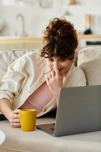 Una joven se relaja en un sofá, tomando café mientras se conecta con su computadora portátil durante la tarde. - foto de stock