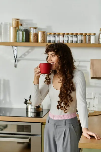 A young woman with curly hair sips from a bright red mug while embracing her kitchen space. — Stock Photo